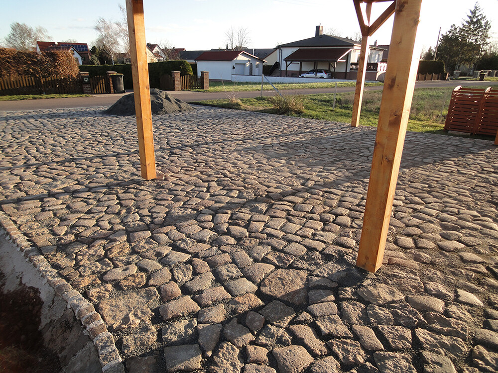 Kopfsteinpflasterfläche mit mehreren Holzbalkenstützen; Bauhaufen im Hintergrund und Häuser in der Ferne.