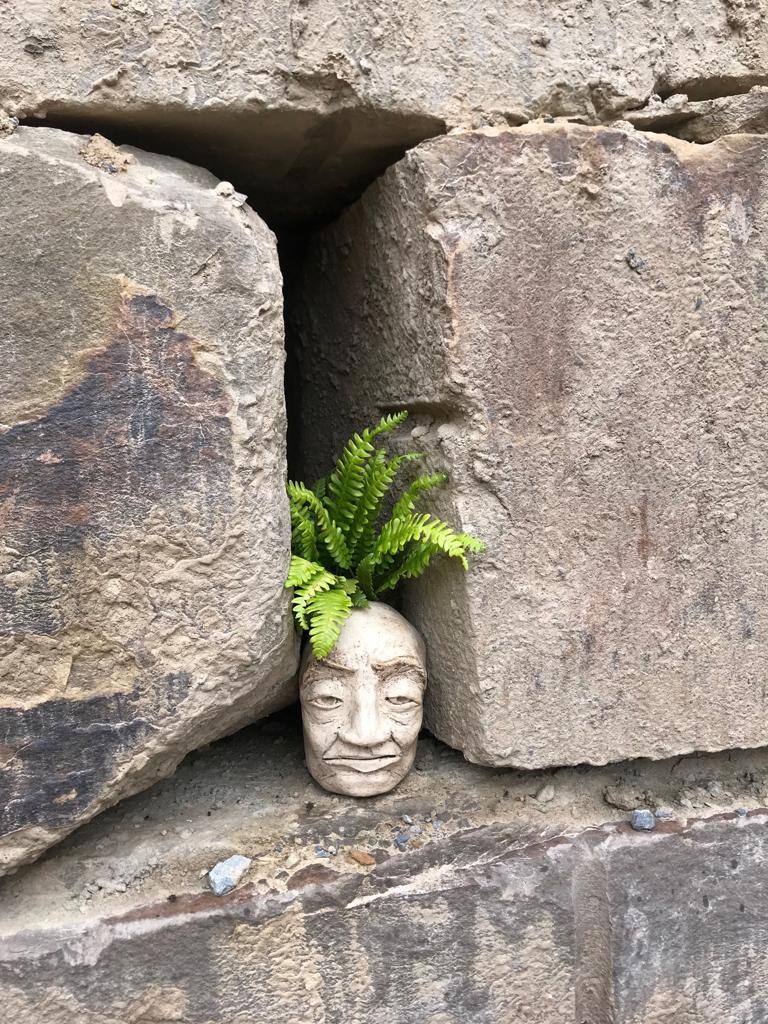 Steinfigur eines menschlichen Kopfes mit Farnen aus dem Oberkopf, zwischen Felsbrocken.
