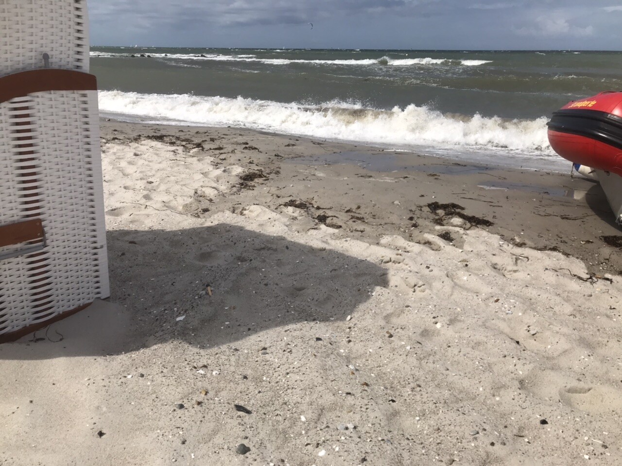 Strand mit Sand, Strandkorb links, brechende Wellen; rotes Boot rechts am Ufer.