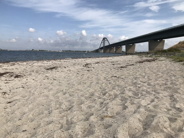 Sandstrand am Ufer mit moderner Brücke über dem Wasser im Hintergrund.