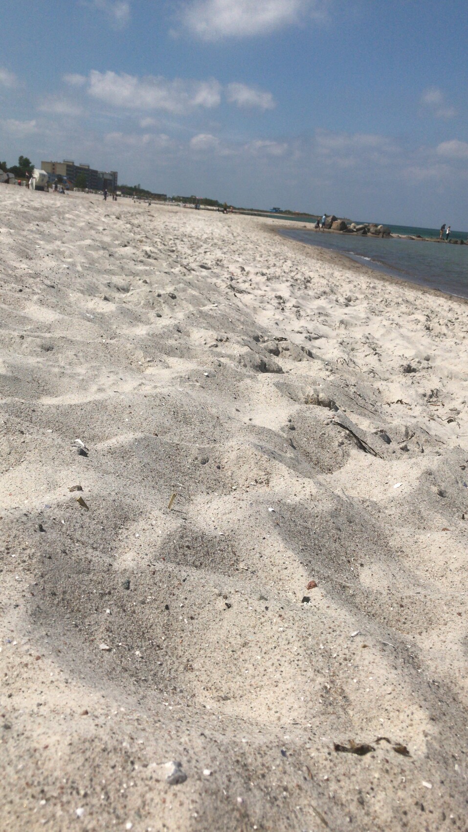 Weitwinkelaufnahme eines sandigen Strandes am Meer mit Wolken im blauen Himmel.