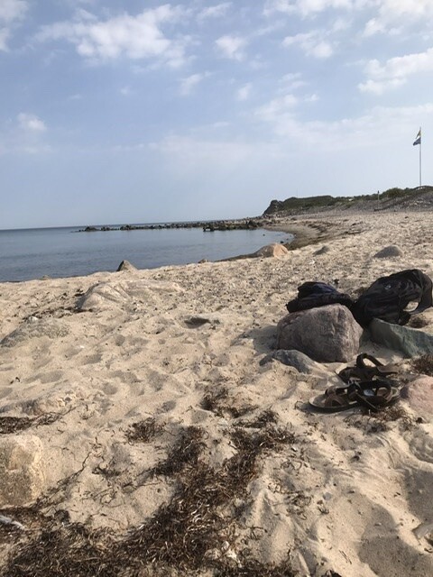 Strand mit Sand, Felsen, Rucksäcke und Schuhe, Meer und Flaggenmast im Hintergrund