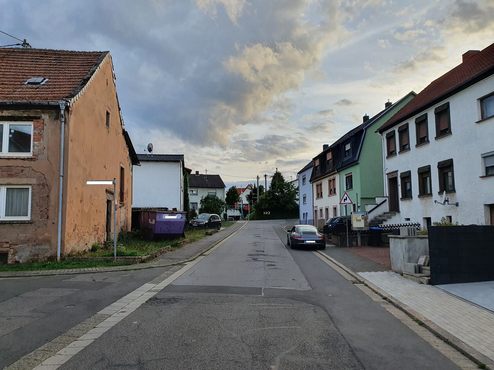 Wohnstraße mit bunten Häusern links und rechts, geparkten Autos, Wolken am Himmel.