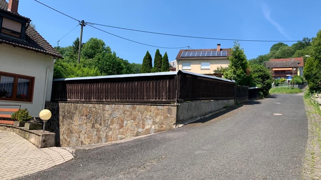 Straße in einem Dorf, Steinmauer mit Holzzaun, links Haus, Häuser am Hang, blauer Himmel.