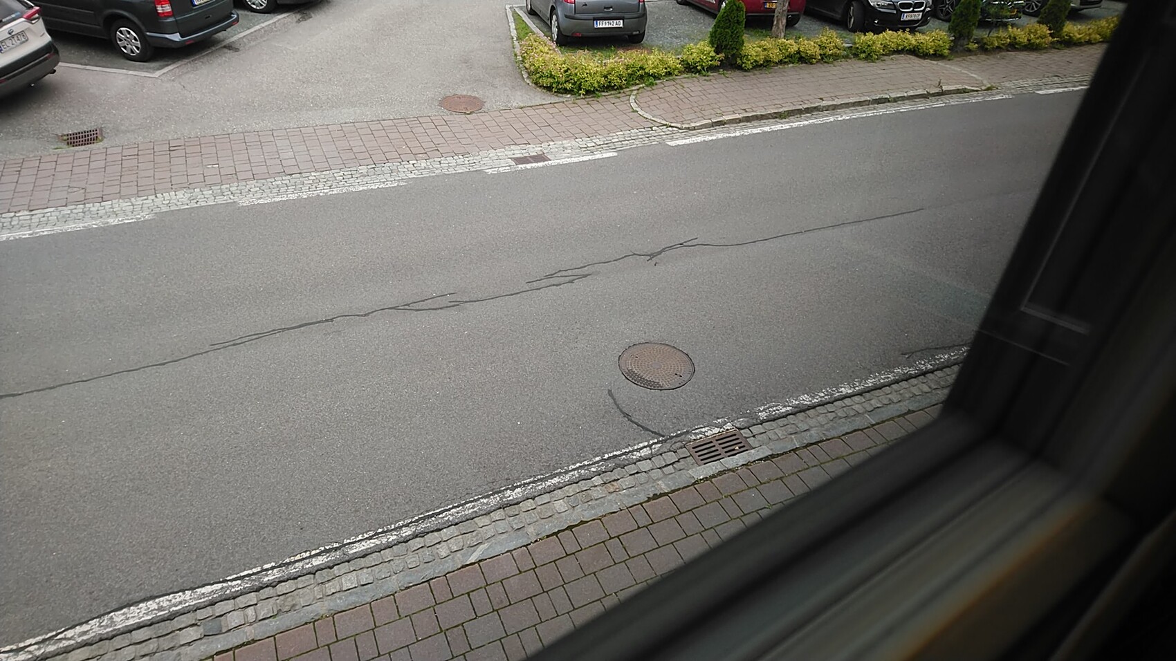 Blick aus dem Fenster auf eine Straße mit geparkten Autos, Gehweg, Kanaldeckel und Rissen im Asphalt.