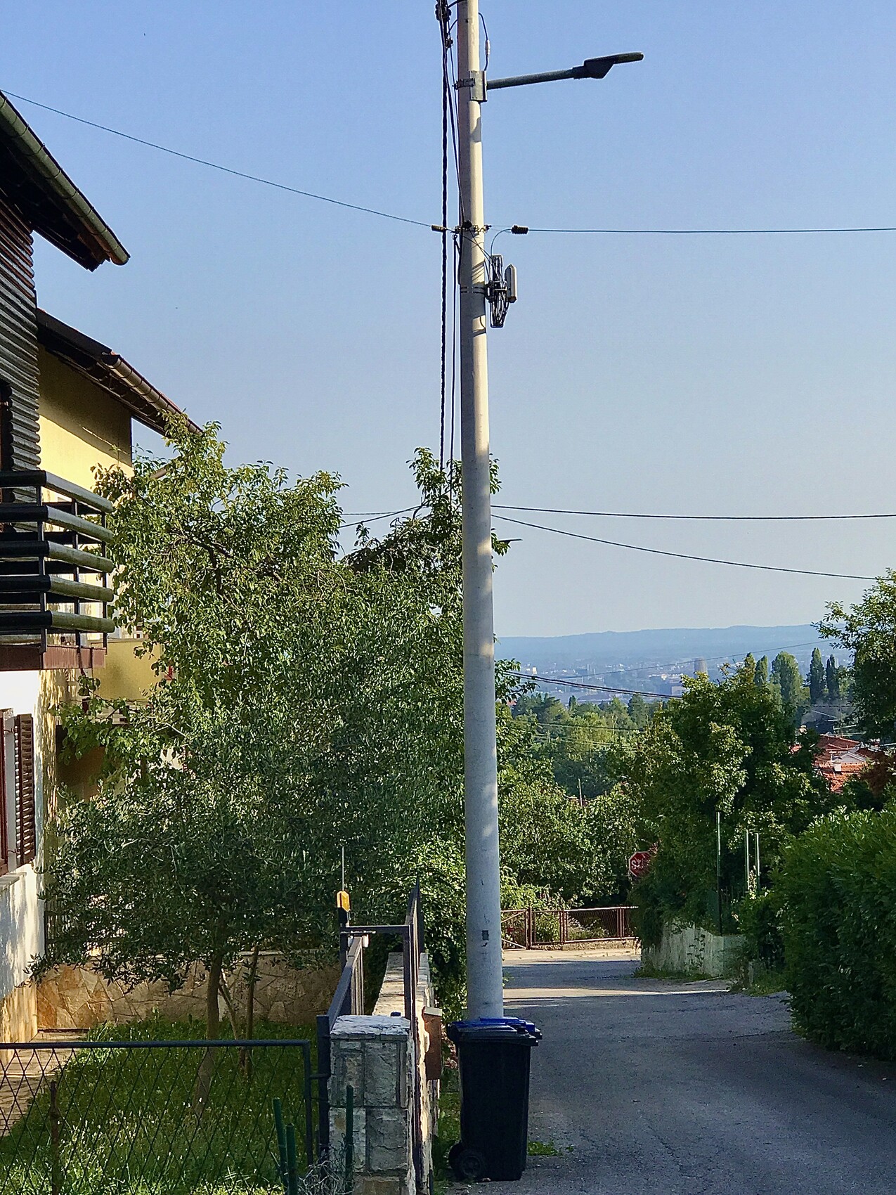 Straßenzug mit Mast, Leitungen und Laterne, links gelbes Haus und grüne Bäume.