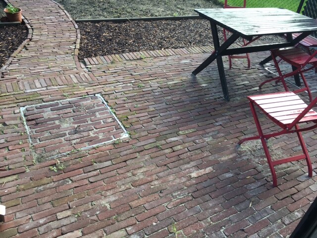 Garten-Patio aus rotem Ziegelboden mit dunklem Tisch und roten Stühlen; Pflanze links.