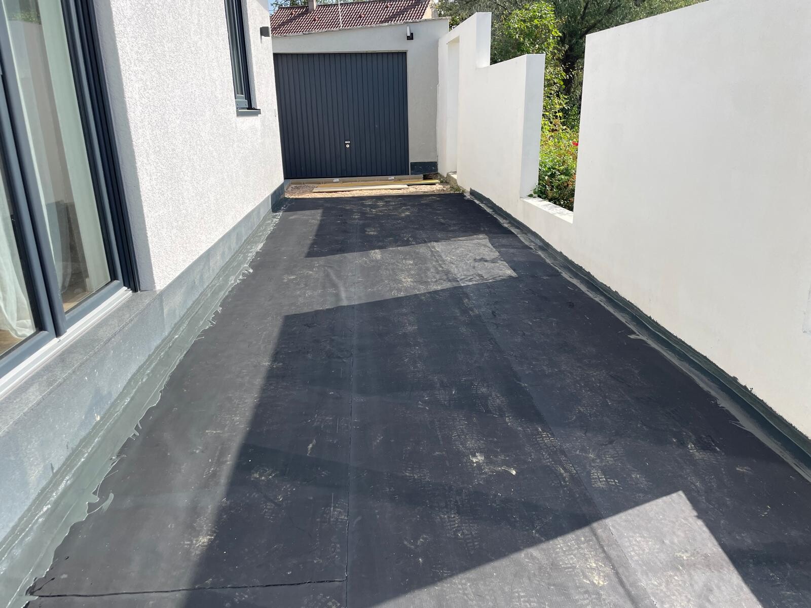 Aussen-Terrasse mit schwarzer Bodenabdichtung, weissen Wanden und Garagentür am Ende