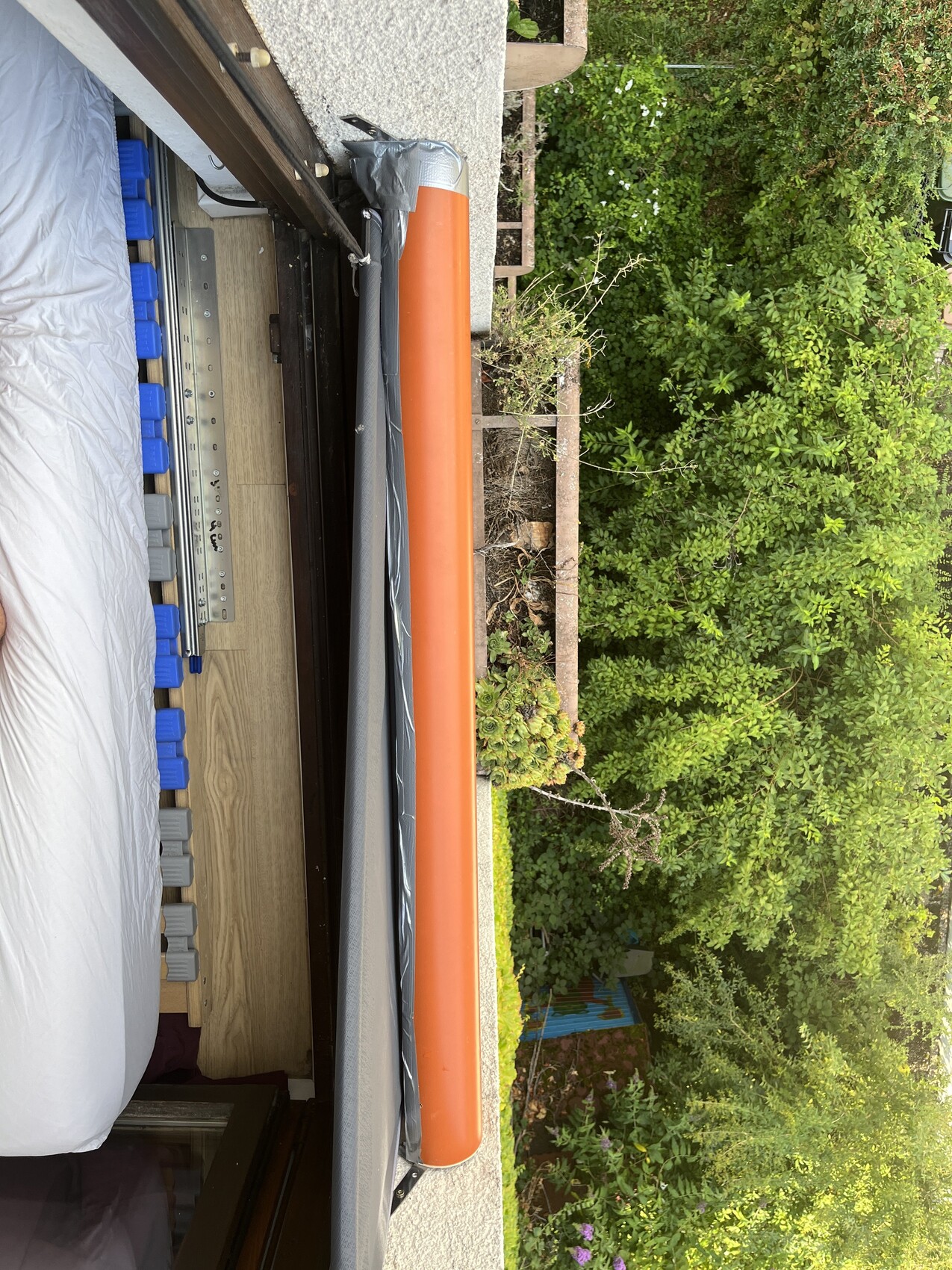 Bett mit Matratze auf Balkon; orangefarbenes Rohr am Geländer, Garten im Hintergrund