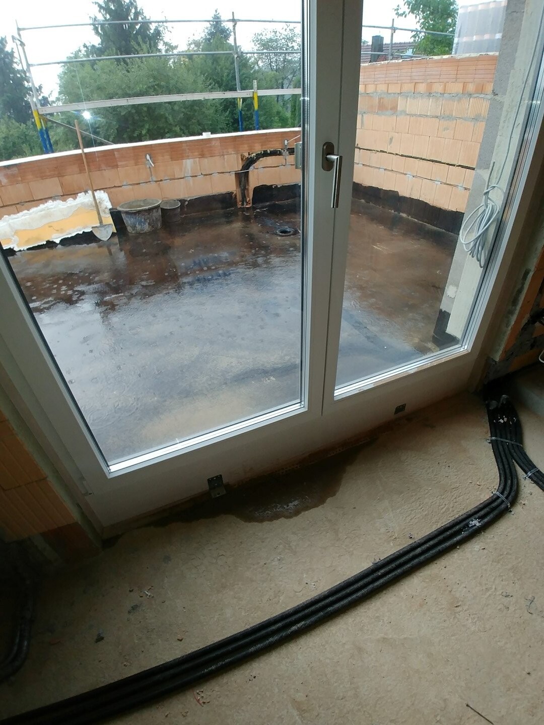 Balkon im Bauzustand mit feuchtem Boden, Glas-Tuer, Außenmauer aus Ziegeln und Gerüst sichtbar.