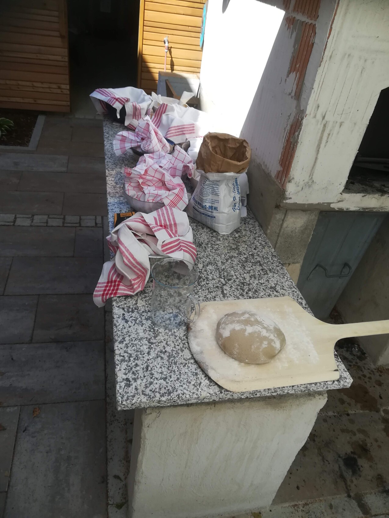 Ein Teigballen liegt auf einem Holzschieber neben Mehlbeuteln auf einer Granitplatte im Freien.