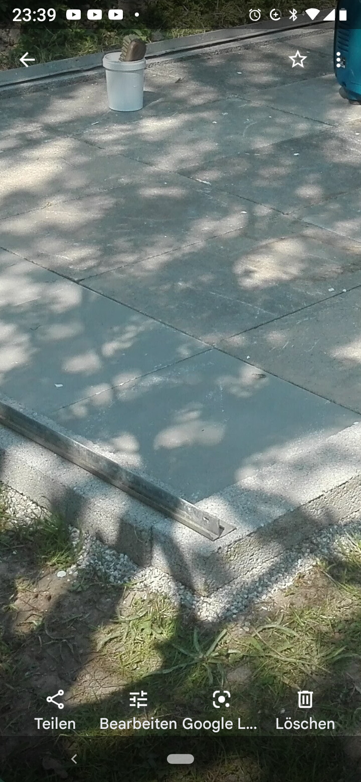 Gepflasterte Steinplatten-Terrasse mit Eimer, Pinsel, Metallstreifen; Schatten von Bäumen.