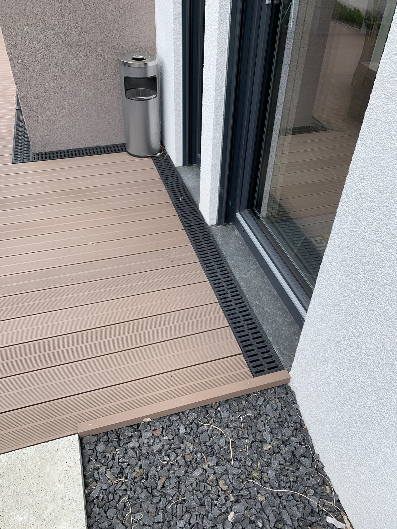 Außenbereich: braune Terrassendielen, Glastür, schwarze Abflussrinne, Metallmülleimer, Kies.