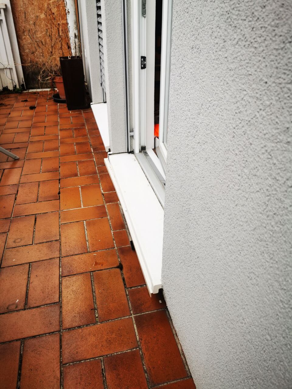 Offene Terrassentür mit weißem Fensterrahmen; orange-braune Fliesen und graue Außenwand.