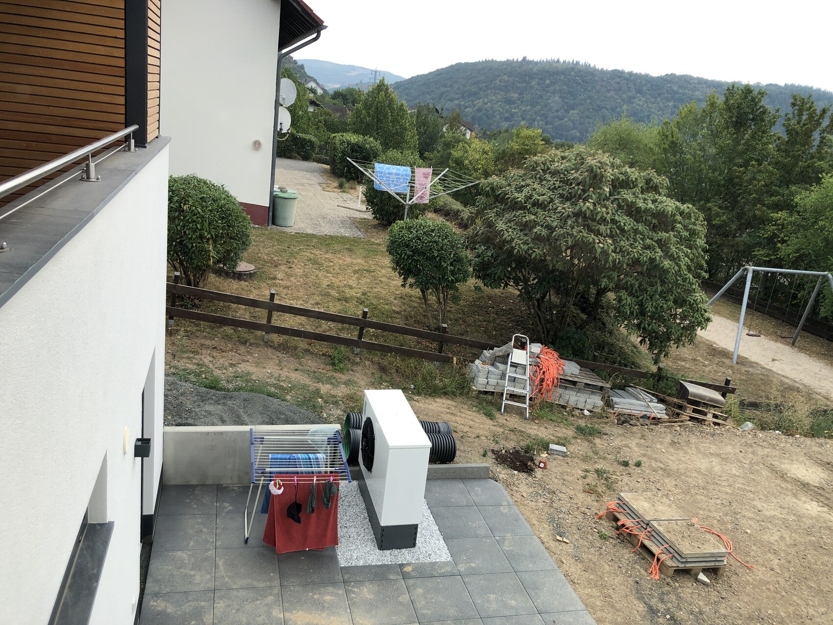 Terrasse eines Hauses mit Wäscheständer, Garten, Bäumen, Spielplatz und Berglandschaft im Hintergrund