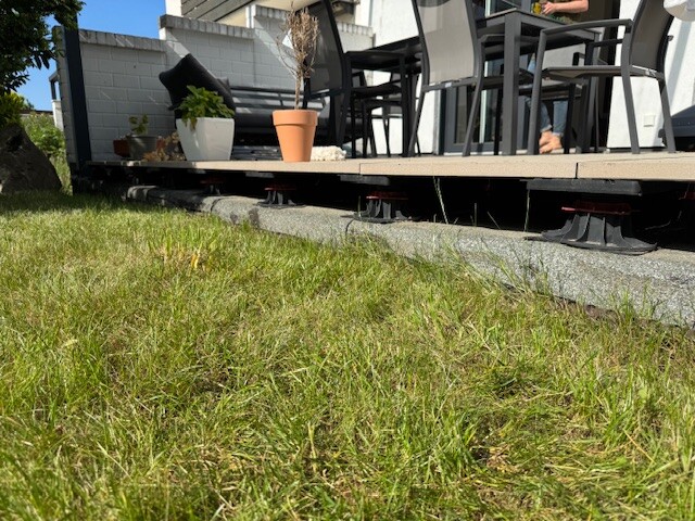 Garten-Terrasse mit Stühlen, Tisch und Topfpflanzen, Gras im Vordergrund.