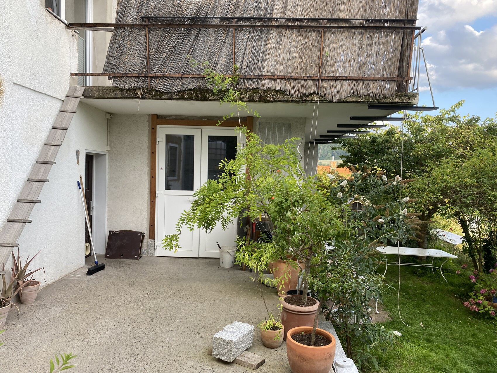 Terrassenbereich vor Haus mit Reetdach-Balkon, weiße Tür, Leiter links, Topfpflanzen