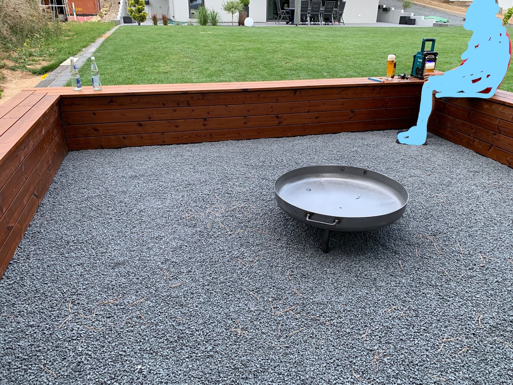 Gartenhof mit runder Feuerstelle auf Kies, umlaufende Holzwände; blaue Umrissfigur rechts.