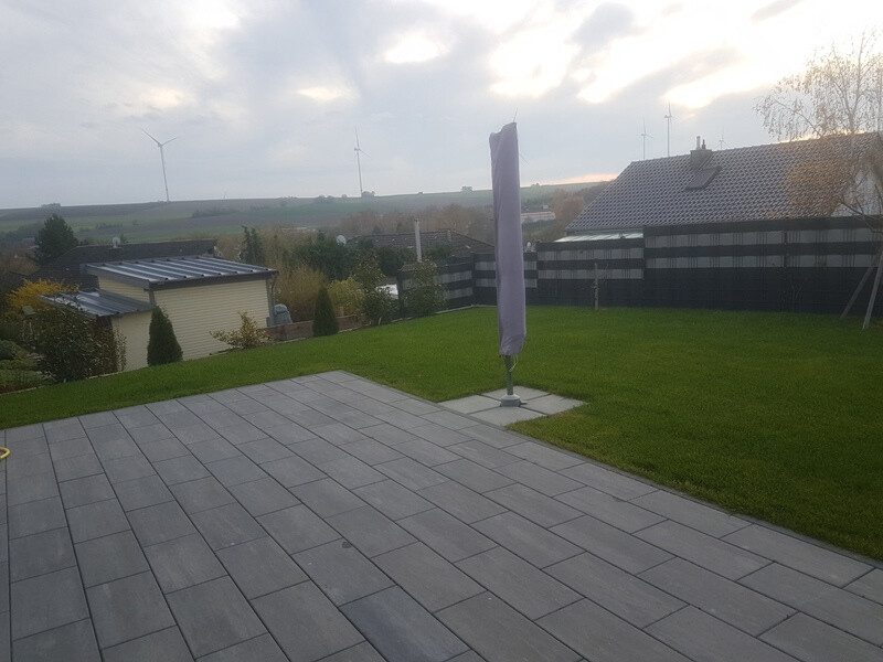 Gepflasterte Terrasse mit geschlossenem Sonnenschirm, Gras und Windräder im Hintergrund.