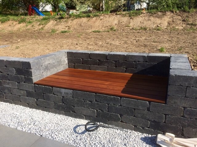 Eingelassene Holzbank in einer dunklen Steinmauer, umgeben von Kies im Gartenbereich.