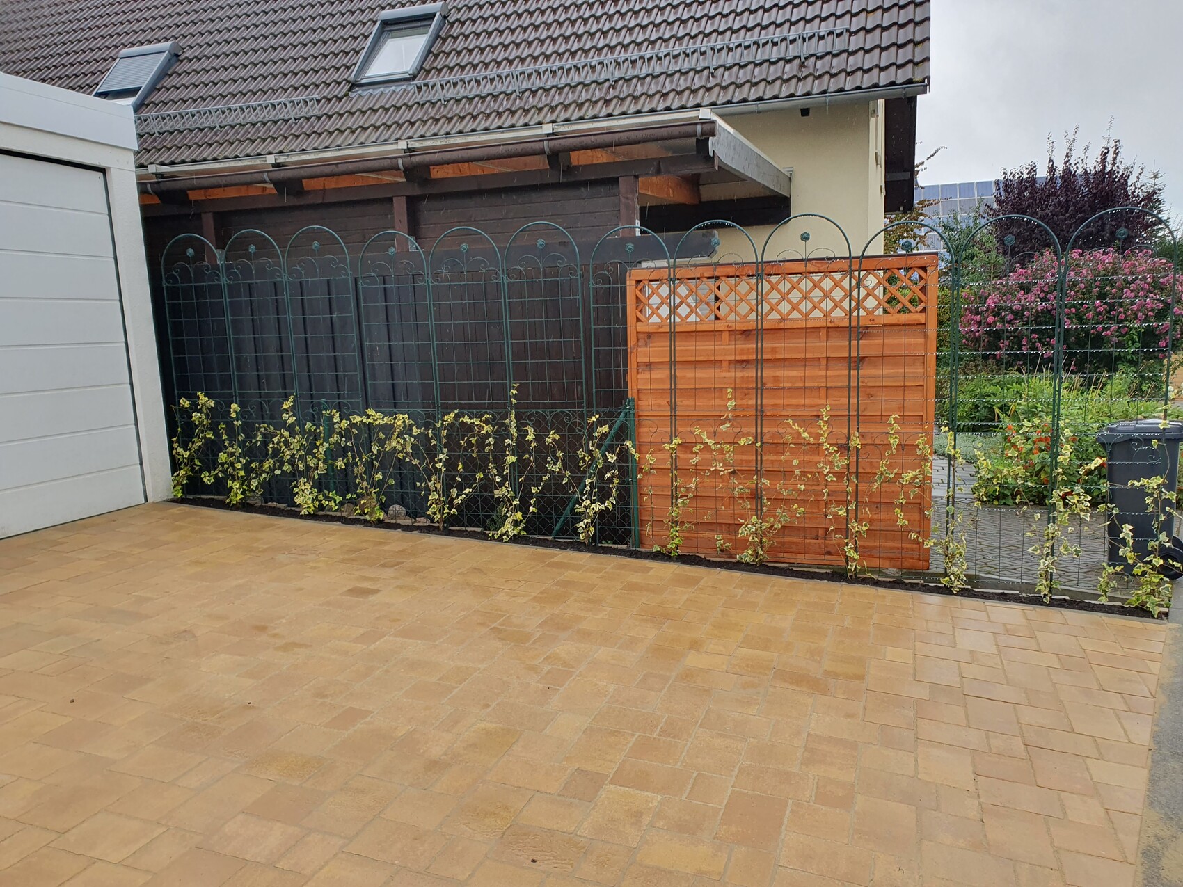 Gepflasterte Terrasse vor Gartenzaun; Metallzaun mit Rundbögen, Holzwand, Kletterpflanzen.