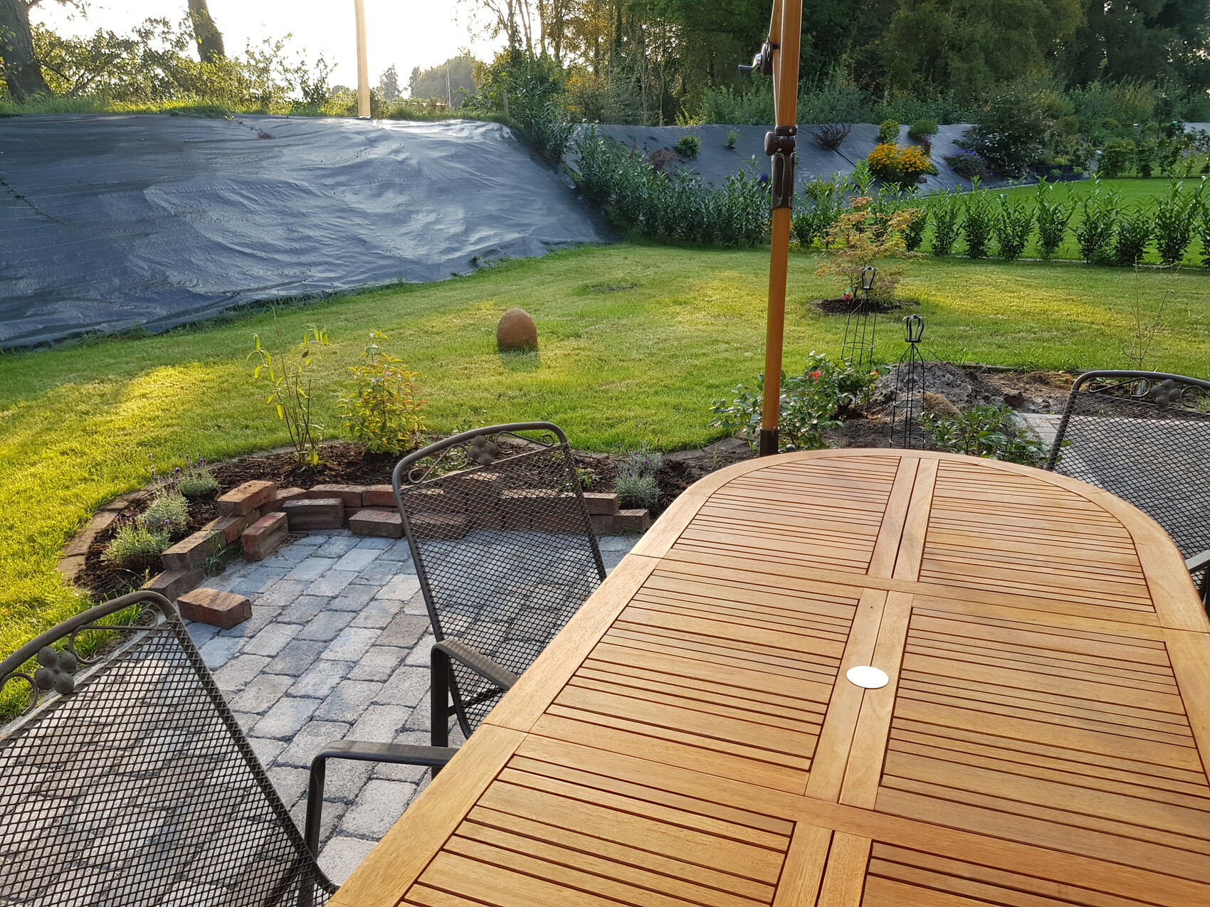 Garten: Großer Holztisch mit Metallstühlen auf Terrasse; grüne Wiese und Beete im Hintergrund