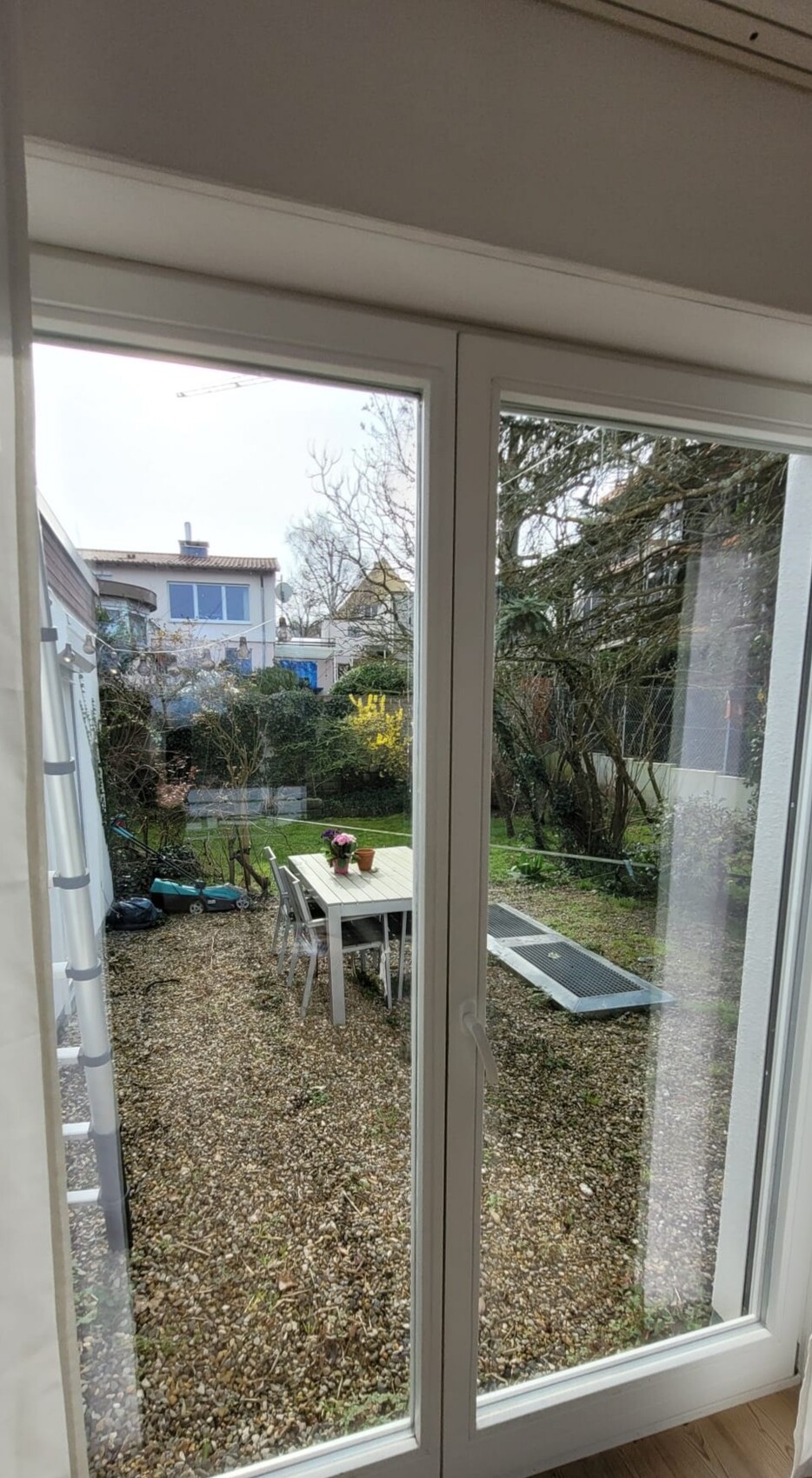 Gartenansicht durch Glas-Tür: weißer Esstisch mit Stühlen, Blumentopf, Kiesboden, Bäume.