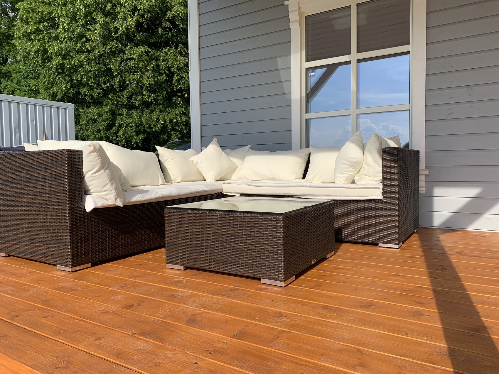 Rattan-Eckcouch mit cremefarbenen Kissen und Glas-Tisch auf Holzterrasse vor Hausfenster.