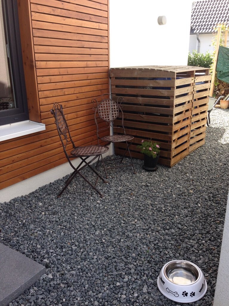 Gartenbereich an Holzfassade mit zwei Metallstühle kleinem Tisch, Pflanze und Hundeschüssel auf Kies