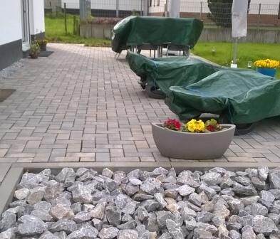 Gepflasterte Terrasse mit Steinplatten, grün abgedeckten Gartenmöbeln und Blumentöpfen.