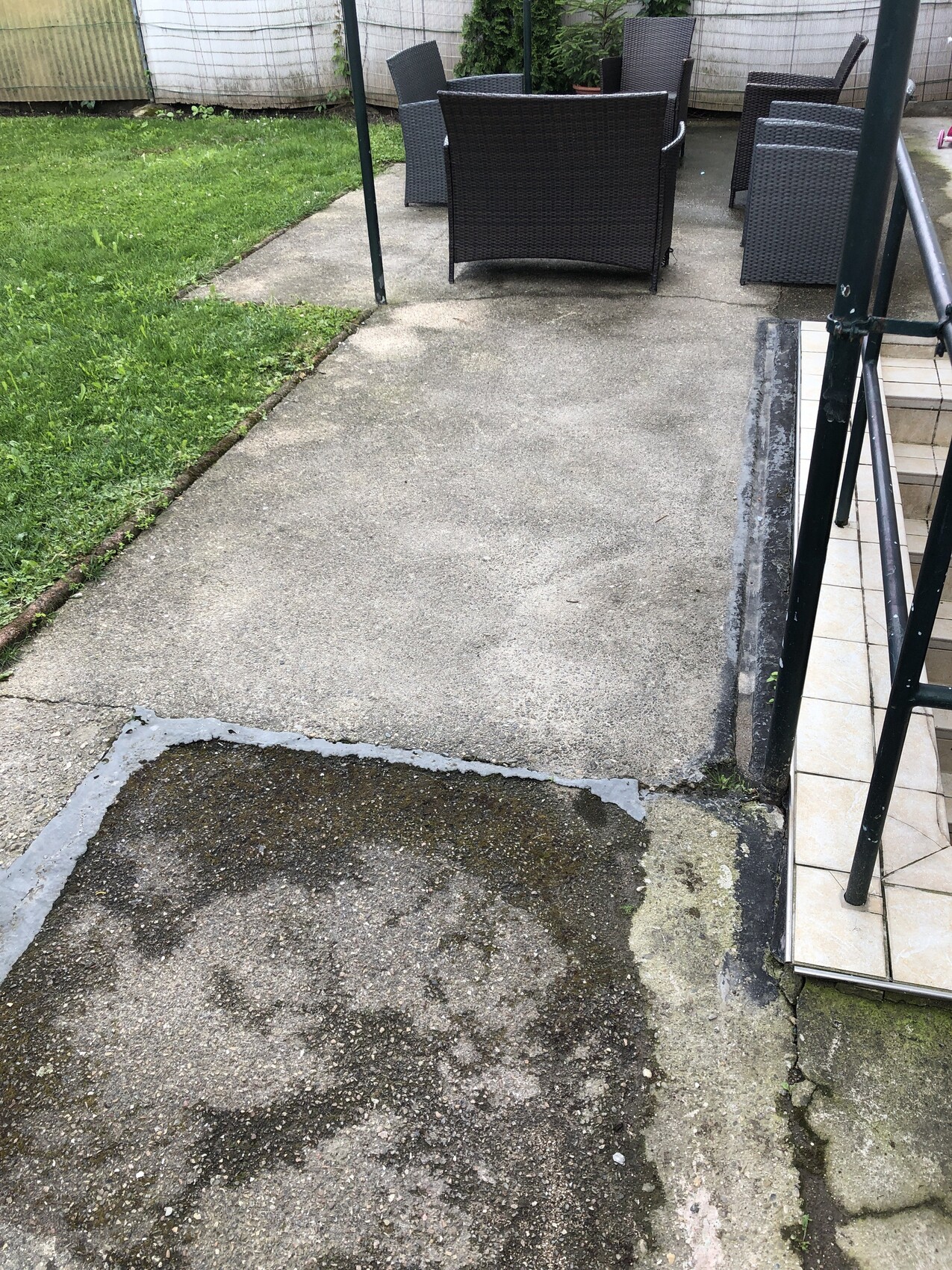 Garten mit dunkler Rattan-Sitzgruppe auf Terrasse; abgenutzter Betonboden und Gras links.