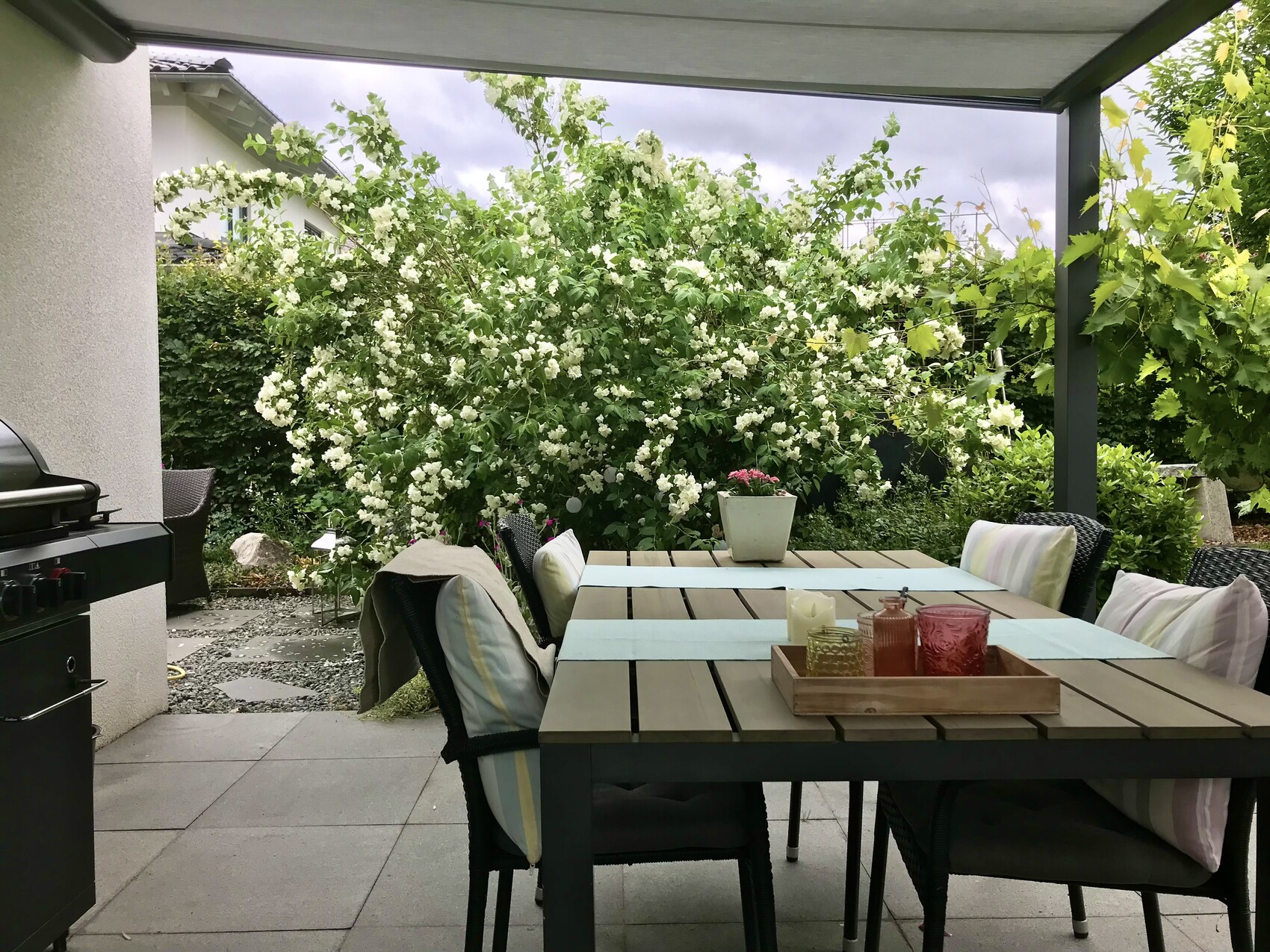 Gedeckter Gartentisch mit Stühlen, Grill links, weißer Blütenstrauch im Hintergrund.