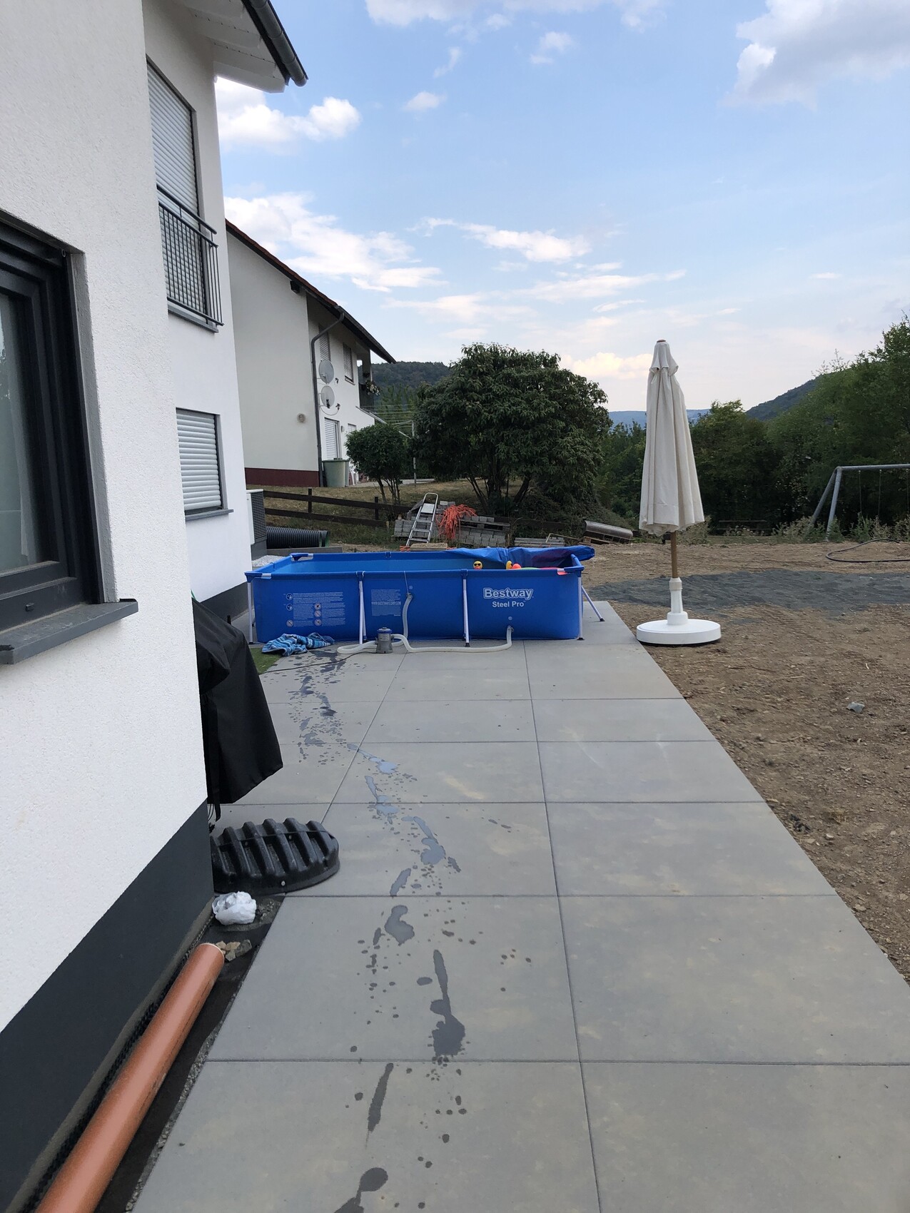 Blaue rechteckige Bestway-Pool auf Terrasse neben Haus; offener Sonnenschirm, Gartenblick und Berge.
