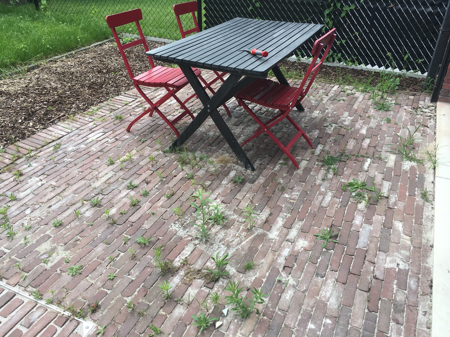 Garten mit schwarzem Tisch und roten Klappstühlen auf Ziegelpflaster; Unkraut sprießt.