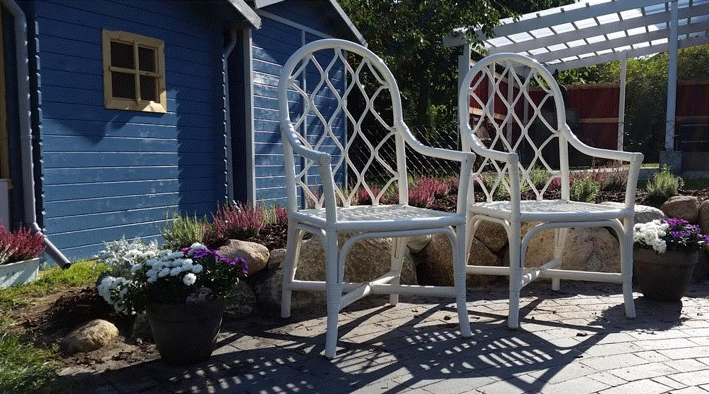 Zwei weiße Gartenstühle mit Gitterrücken stehen auf Steinplatten neben Blumen vor blauem Holzhaus.