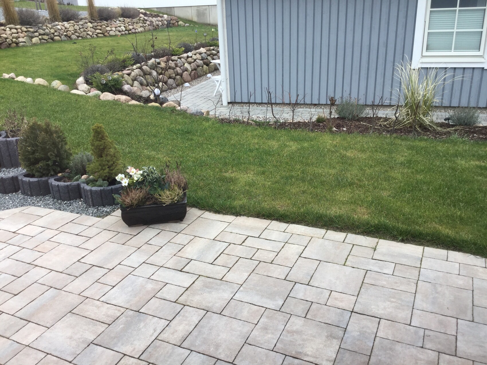 Gepflasterte Terrasse mit rechteckigen Steinen, grünem Rasen und Beetpflanzen am Haus.