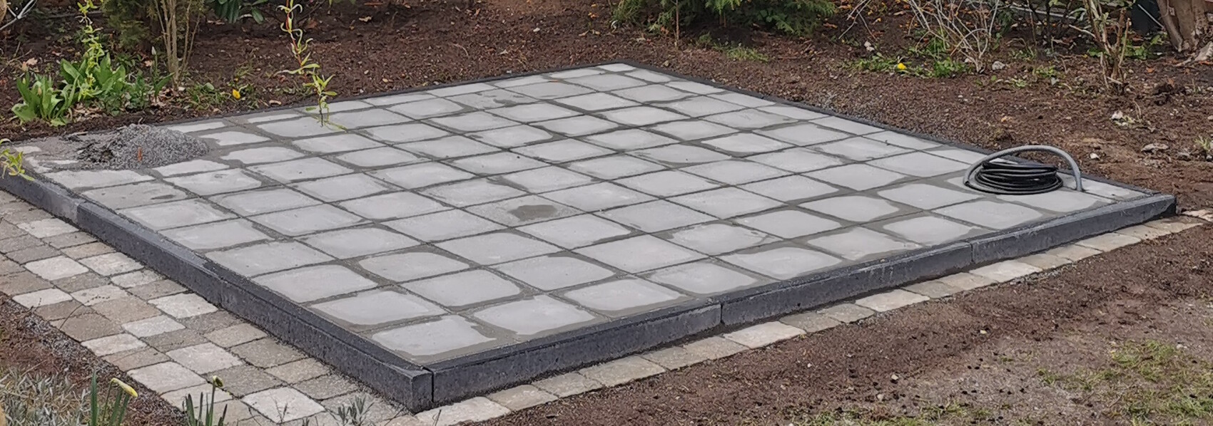 Gepflasterte Terrasse aus quadratischen Pflastersteinen im Garten; rechts eine Kabelrolle.