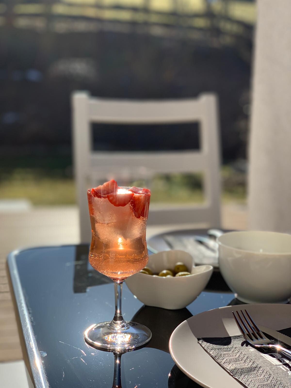 Rosé-Getränk mit Eis und Erdbeeren im Weinglas auf Glastisch im Freien; Oliven-Schale daneben.