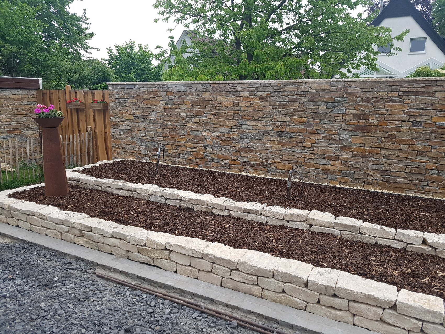 Gepflegter Garten mit zwei erhöhten Steinbeeten, Mulchboden und Steinmauer im Hintergrund.