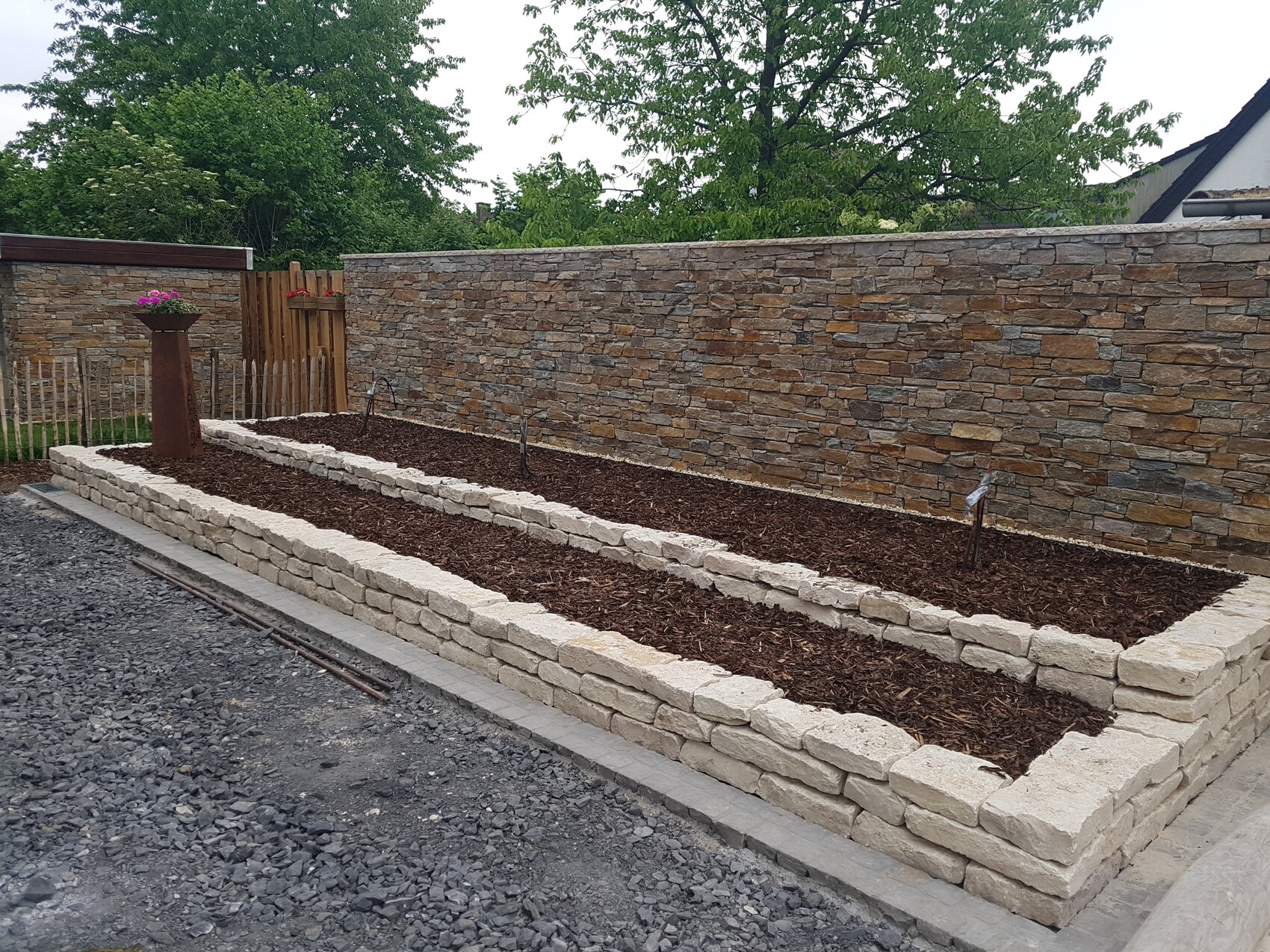 Gartenbereich mit Steinmauer, Mulchbeeten, Pflanzgefäß und Holzzaun im Hintergrund