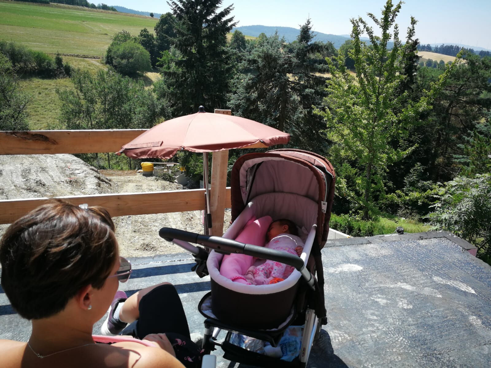 Frau mit Kinderwagen, schlafendes Baby in pinkem Schlafsack, unter pinkem Sonnenschirm auf Terrasse.