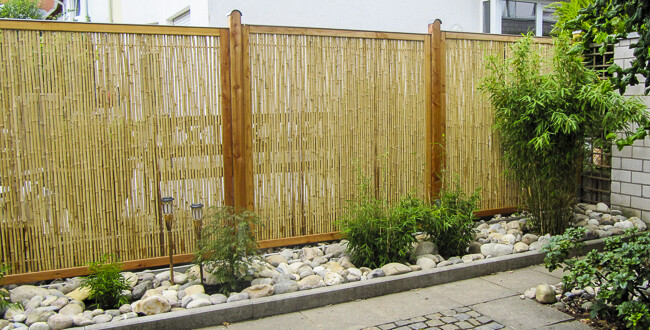 Garten mit Bambus-Sichtschutzzaun, Holzpfosten, Kies und grünem Gebüsch vor einem Pflasterweg.