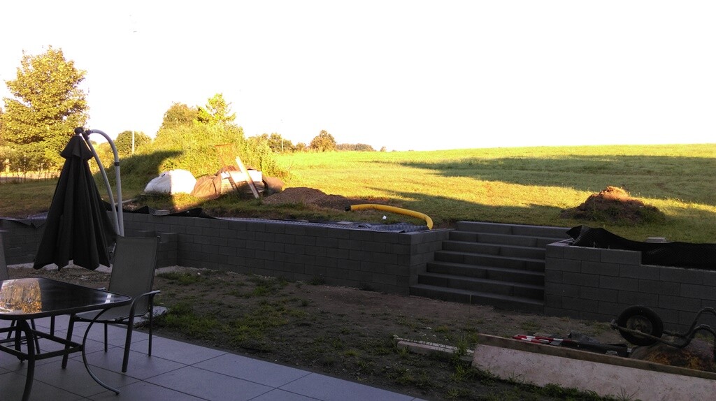 Garten-Terrasse im Bau mit Steinwall, Treppen, Tischgruppe, Sonnenschirm und Baumaterialien.