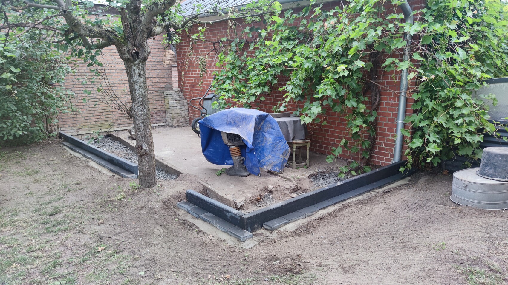 Garten mit Baum, Kletterpflanzen an Backsteinwand, blau abgedeckte Maschine neben Tisch.