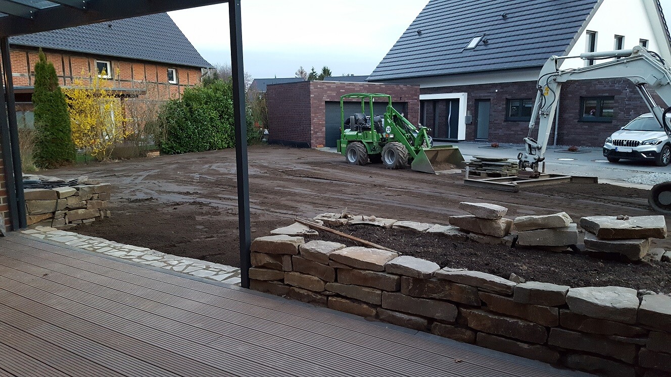Baustelle mit Steinmauerbau, grünem Kompaktlader, Minibagger, Holzdeck und Haus im Hintergrund.