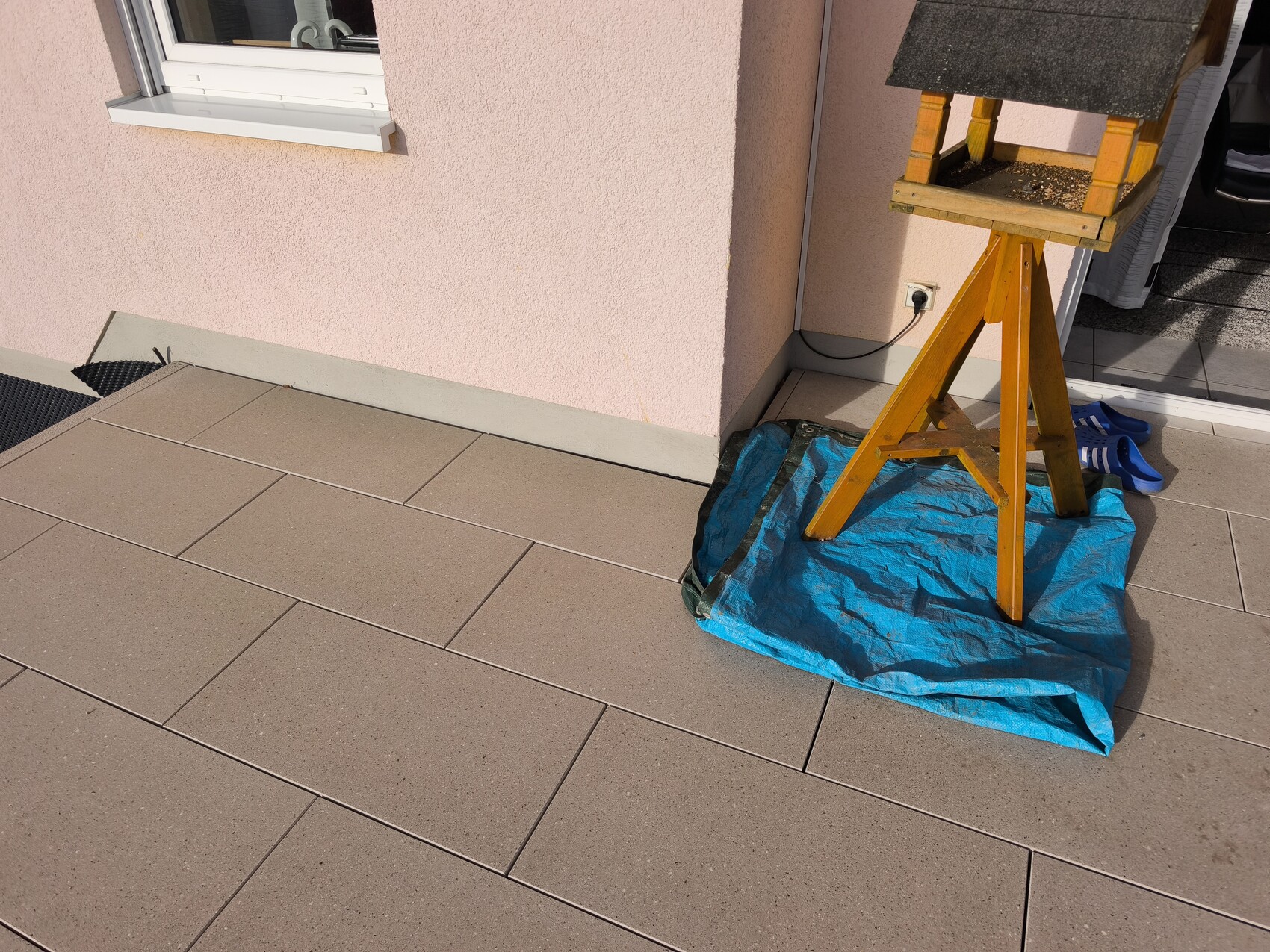 Außenbereich mit Terrassenplatten, pinker Hauswand, Holzständer auf blauer Plane und Schuhen daneben