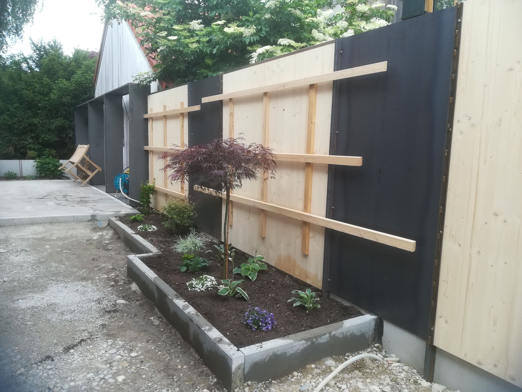 Garten mit provisorischem Lattenzaun, Pflanzbeet und Liegestuhl im Hintergrund.