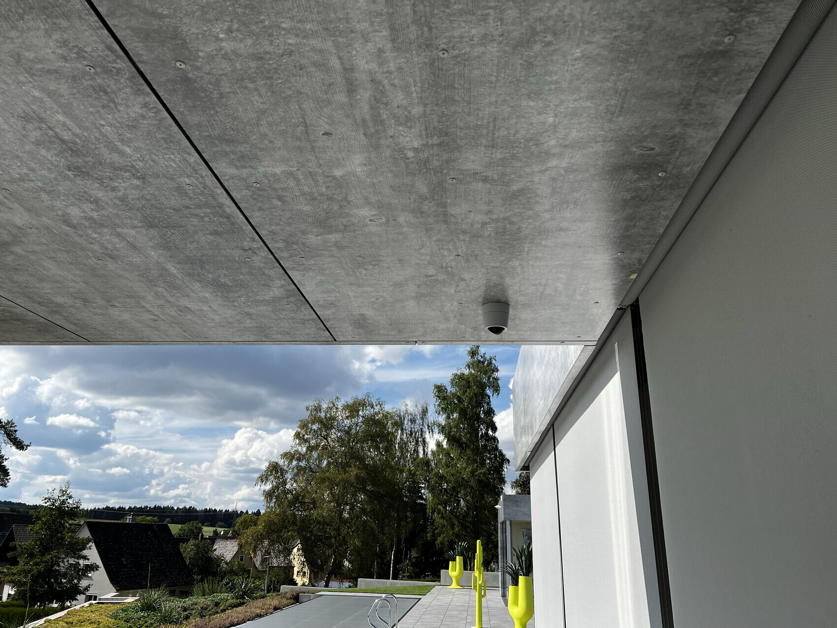 Außenbereich einer modernen Terrasse mit Beton-Decke, Blick auf Garten und Himmel.