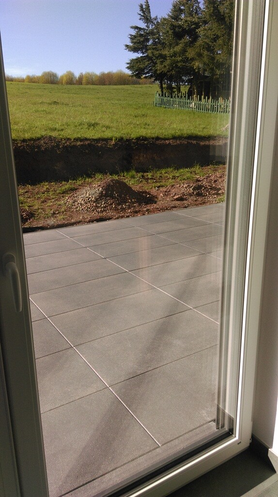 Blick durch Glastür auf Terrasse mit grauen Fliesen, grünem Gras und Bäumen