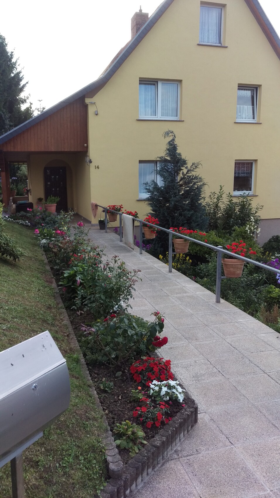 Gelbes Haus mit gepflastertem Weg, blühender Garten am Eingang und Blumenkübel am Geländer.